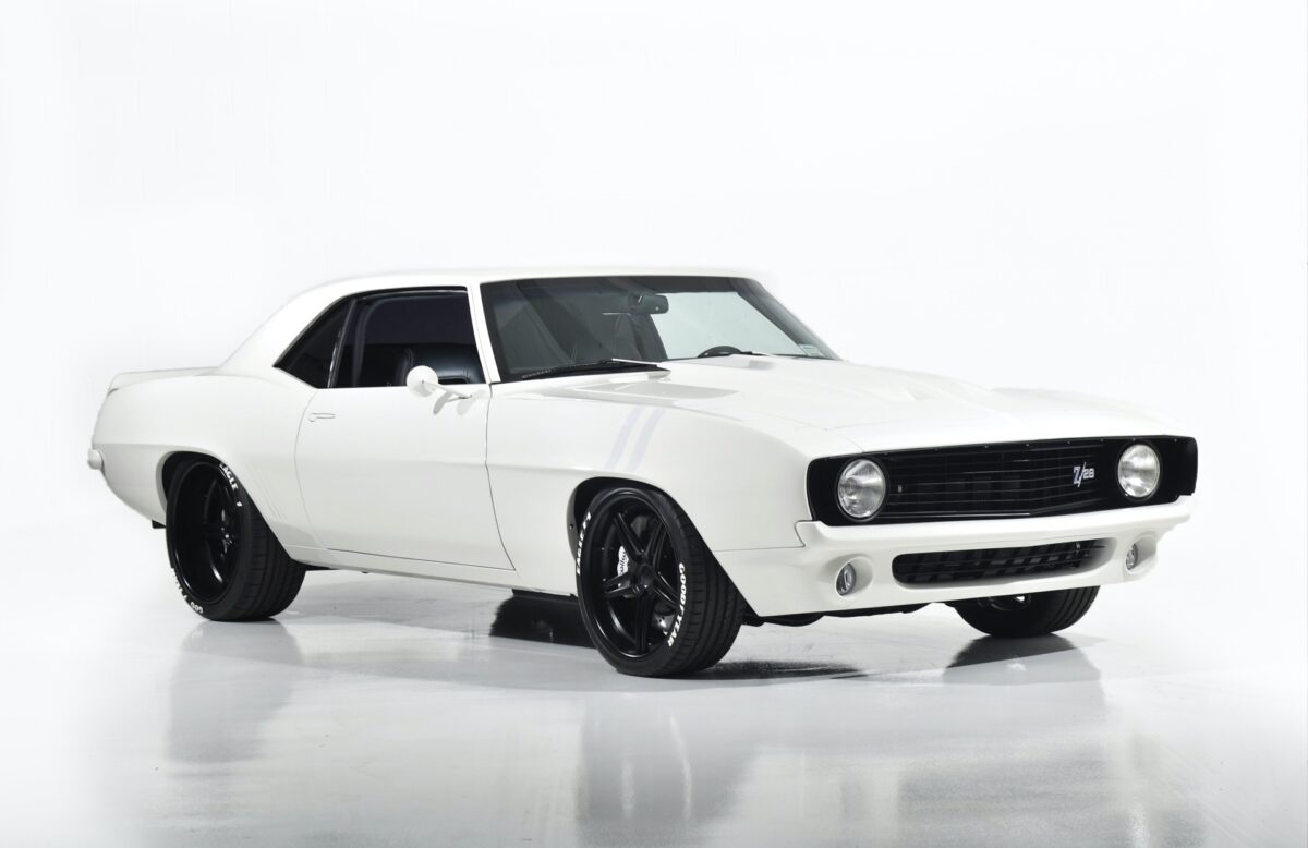 A white vintage Chevy Camaro is parked in an all white garage with a gloss floor.