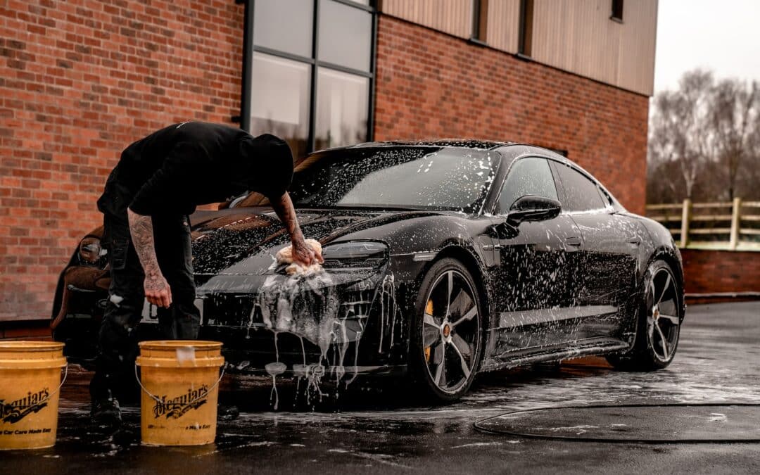 Someone is washing a black Tesla with a hose and soap outside a brick building.