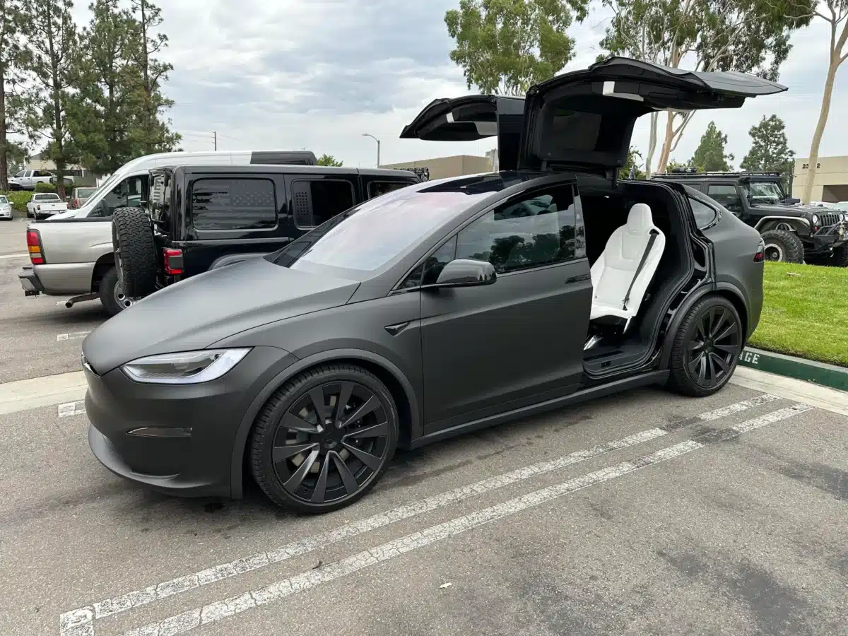 Parked in a parking lot, with the back passenger doors open, there is a Matte Black Tesla Model X.