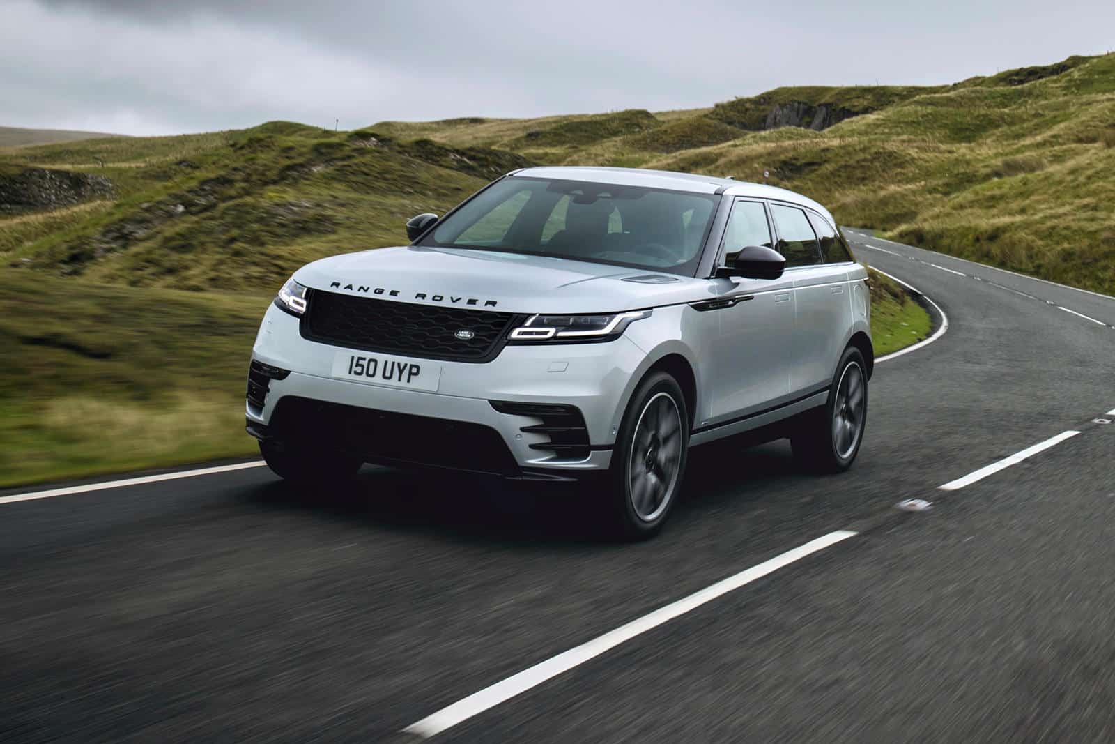 A white range rover velar driving across a paved road in the green hills of the northern united kingdom.