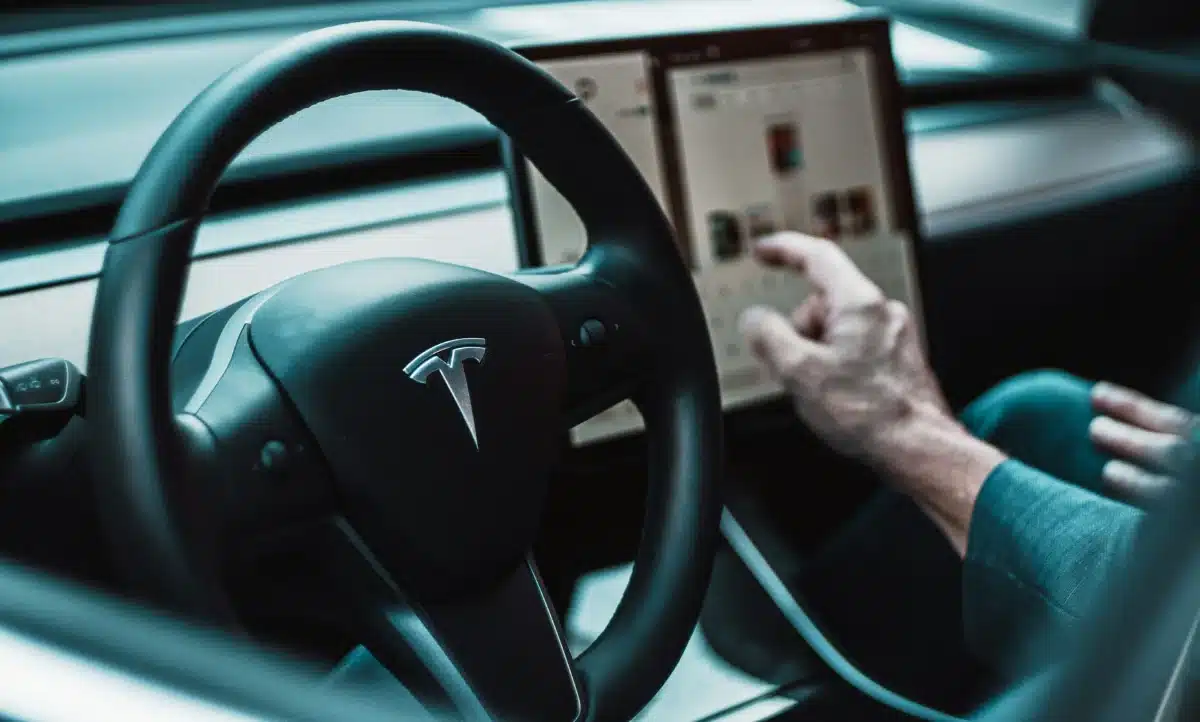 A person is sitting inside the driver's seat of a Tesla. They are pointing at something on the touch screen. Their interior is clean, as if they got a Tesla interior detailing completed. The leather seats are dust free and the windows don't have a single fingerprint.