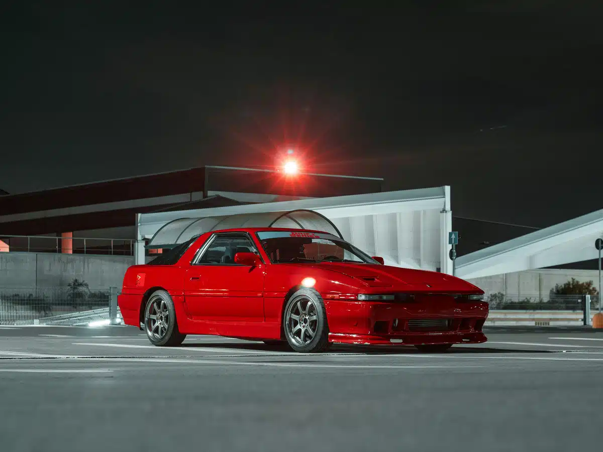 A red Toyota Supra MK3 is parked in a parking lot outside at night.