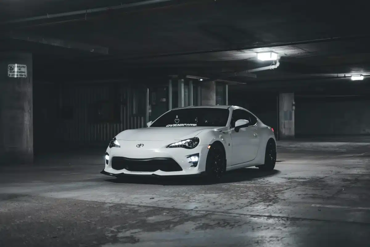 A white Toyota GR86 is parked in a garage with dim lightening and cement flooring.