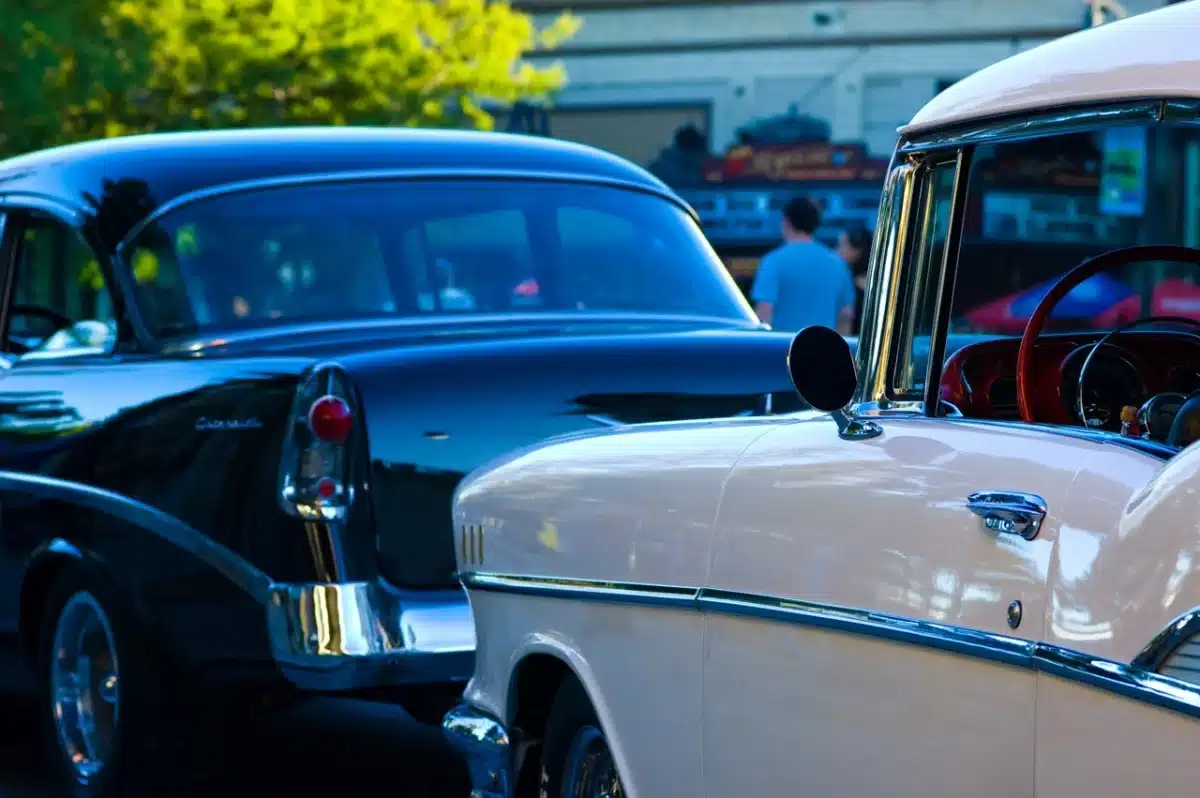 Black and white classic American Graffiti cars are parked on a street outside.