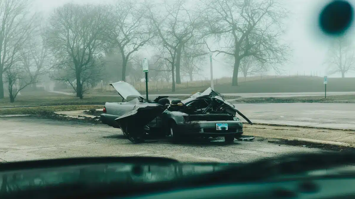 A photo is taken of a damaged car on the side of the road, viewed from the front window of another car. It looks like someone is wondering what to do when someone hits your parked car.