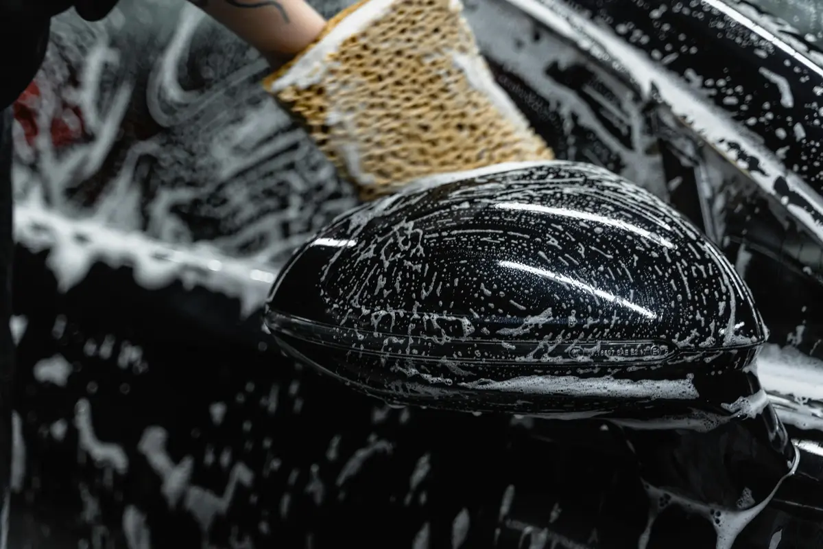 A black vehicle is pictured close up as someone sponges soap or cleaning product over the exterior. 