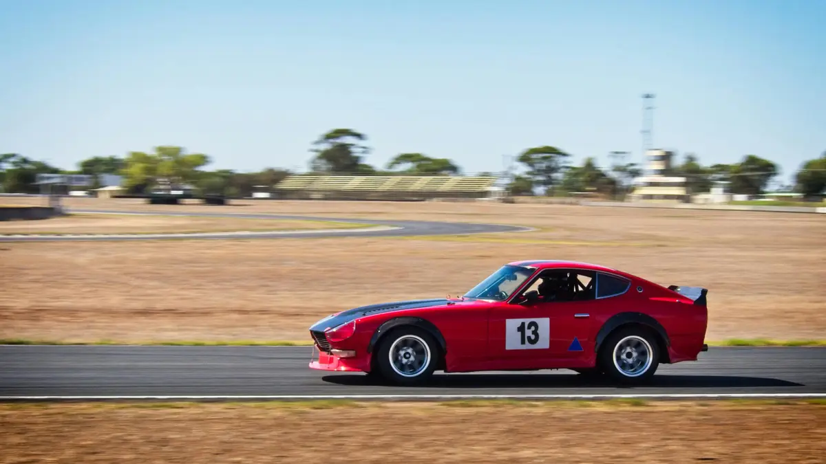 A red race car is pictured on a track in a dry, sandy field. It has the number 13 on the side of the door.