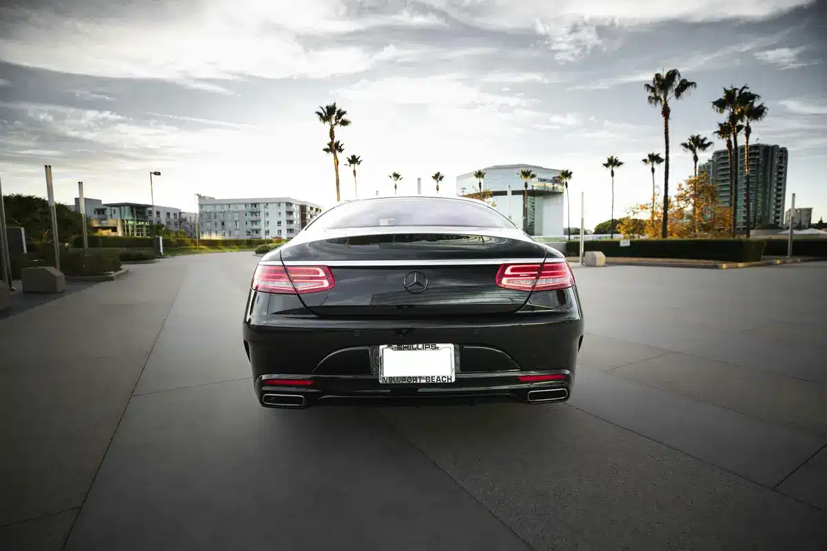 A black car is driving away from the camera on smooth black pavement. There is a cloudy sky with palm trees in the background.