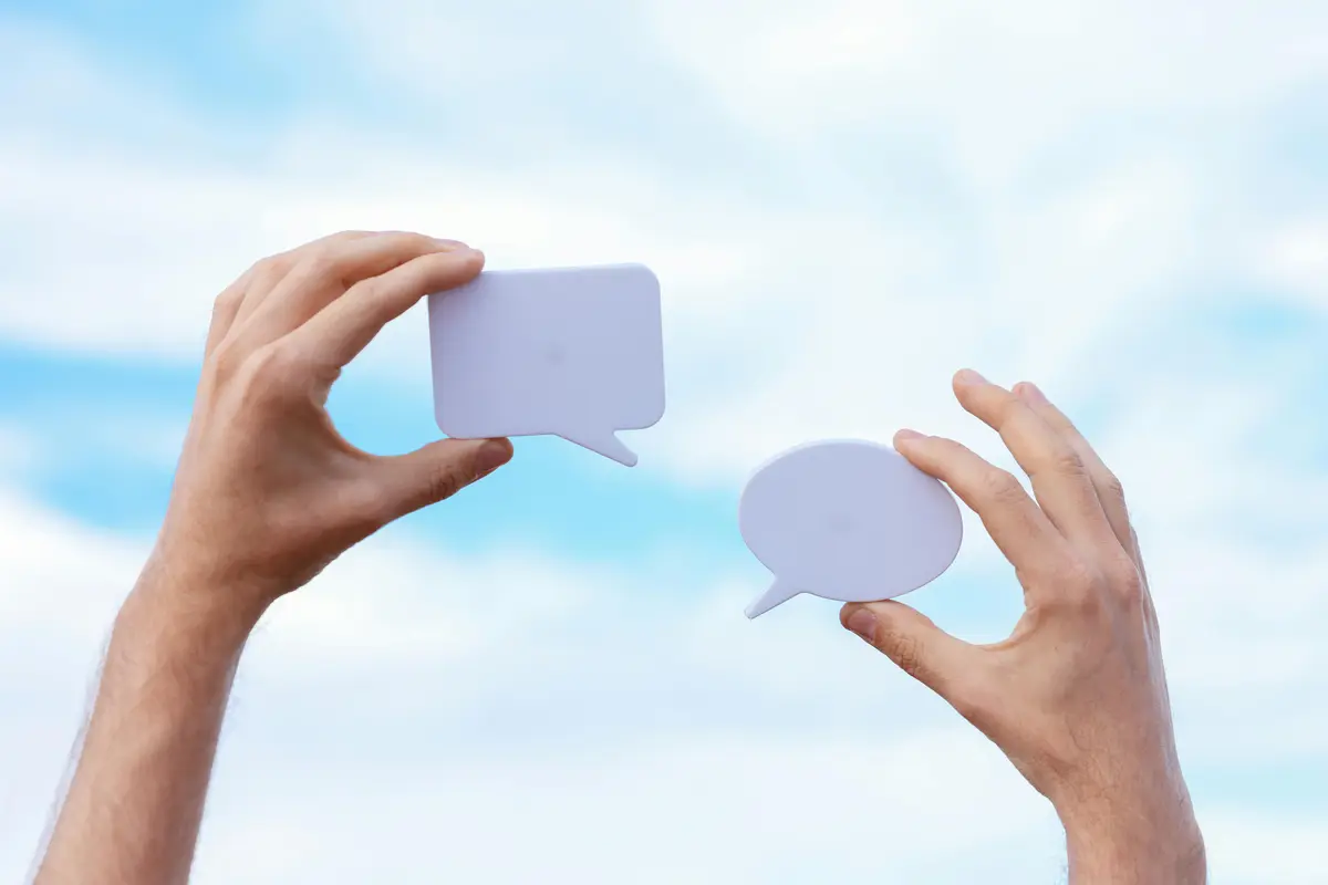 Two hands each holding up one light-blue paper cut-out of a speech bubble. There is a blue cloudy sky in the background.