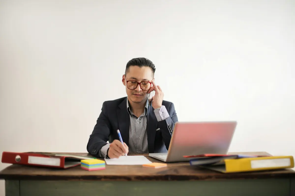 A man is taking a note in a notepad in front of a laptop at a desk. He is also making a phone call.