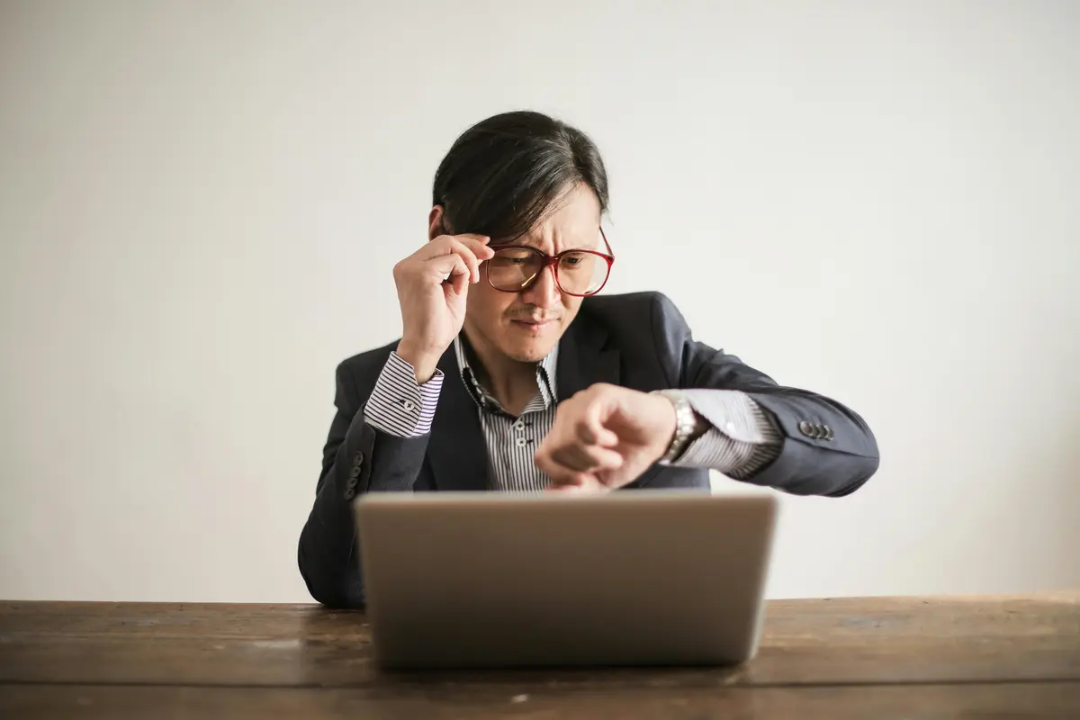 A man is on a laptop at a desk, looking at a watch on his wrist as he squints though his glasses.