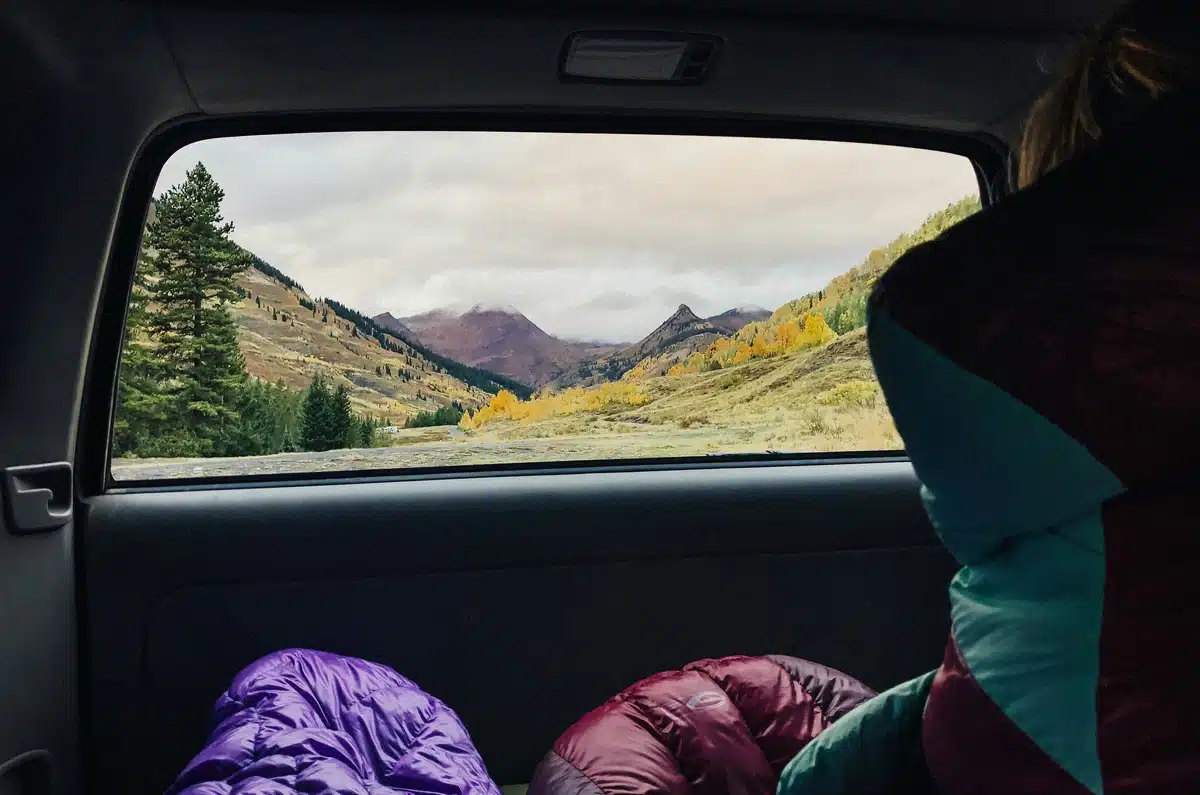 Two sleeping bags are packing in the back trunk of a large SUV, with tinted windows facing the sunny, hilly road behind them.