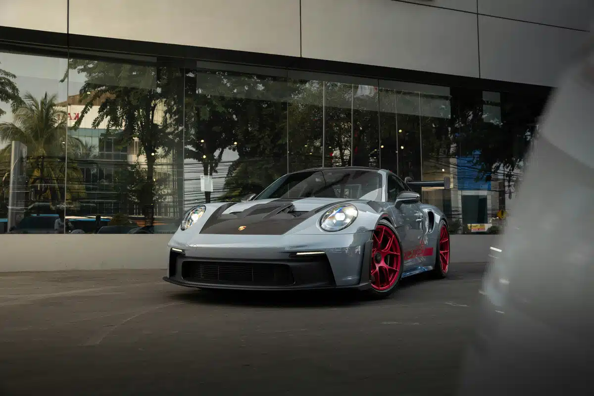 A grey Porsche 911 with a black hood is parked in front of a modern building, boasting a shiny exterior under a shaded parking spot.