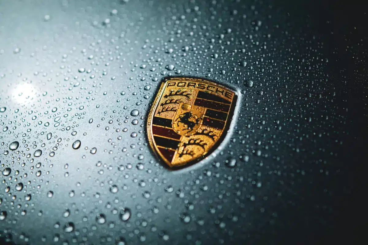 A close-up of a Porsche logo with clean lines around the edge, water drops across the top of the exterior. 