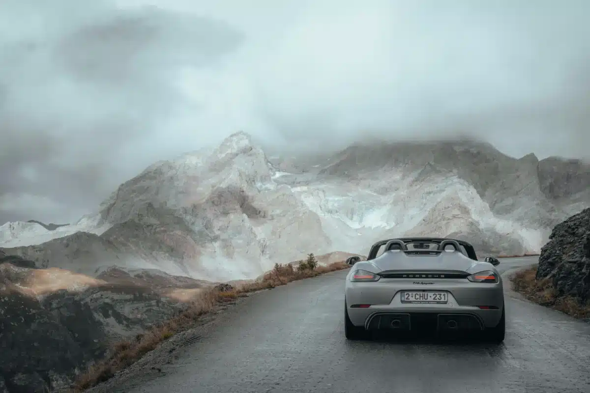 A grey Porsche is being driven on a paved road along a foggy mountain range. 